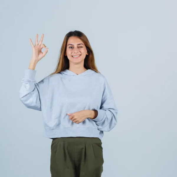 blonde-girl-showing-ok-sign-olive-green-blue-sweatshirt-pants-looking-radiant-front-view